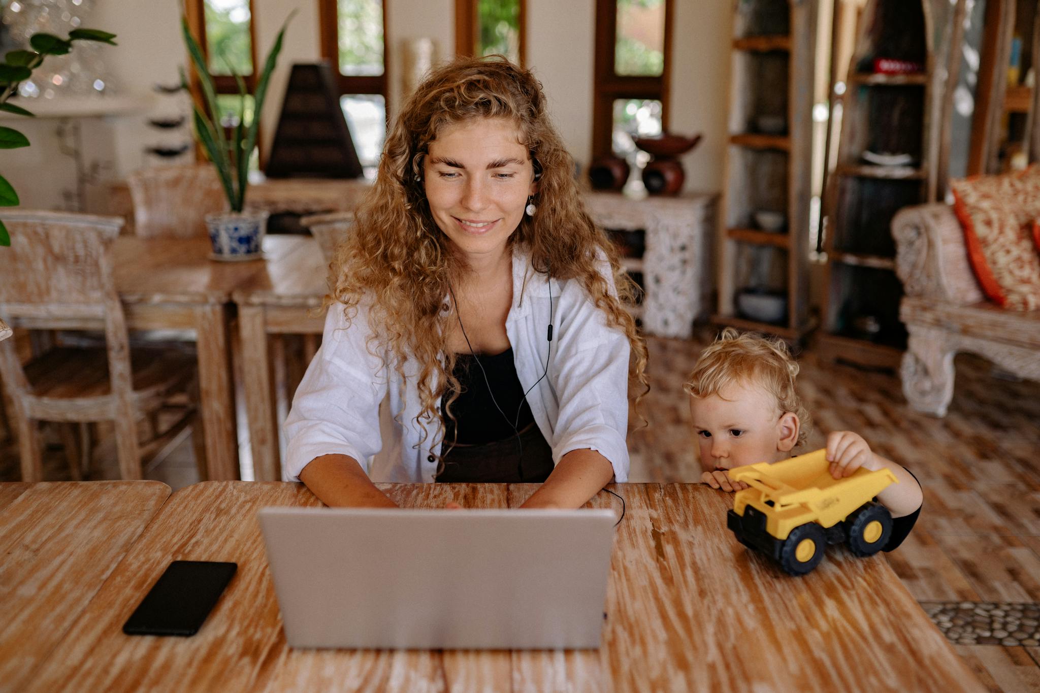 A woman working on a laptop at home with her child playing beside her, creating a balanced family lifestyle.